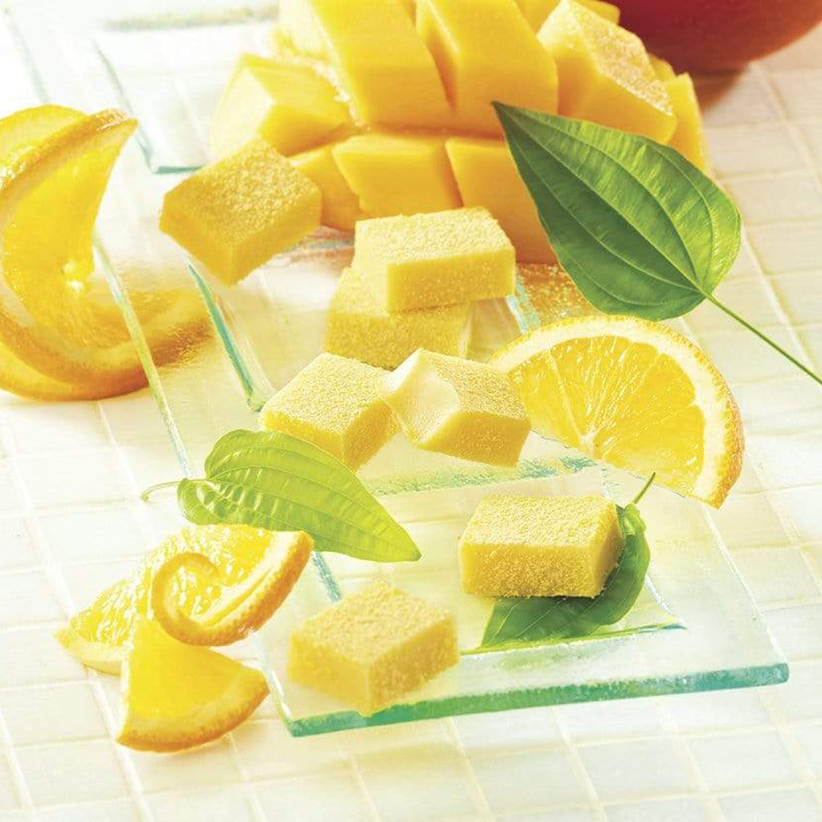 ROYCE' Chocolate - Nama Chocolate "Orange & Mango" - Image shows yellow blocks of chocolate on a clear plate with fruits like orange and mangoes with accents of green leaves. Background features white tiles.