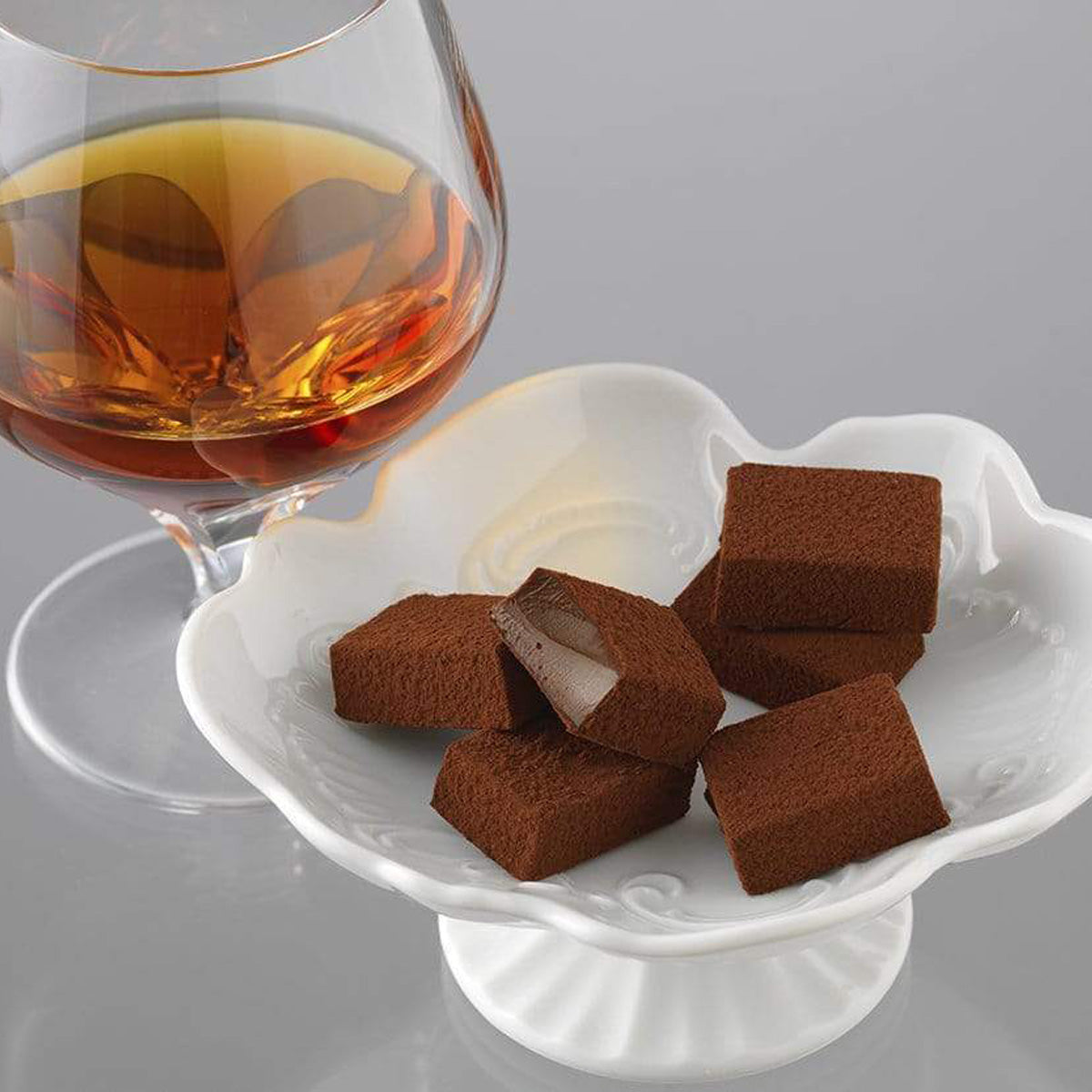ROYCE' Chocolate - Nama Chocolate "Islay Whisky (Port Charlotte)" - Image shows brown blocks of chocolates in a white flower-shaped plate. Also seen is a clear glass on left with amber-hued whisky. Background is light gray.
