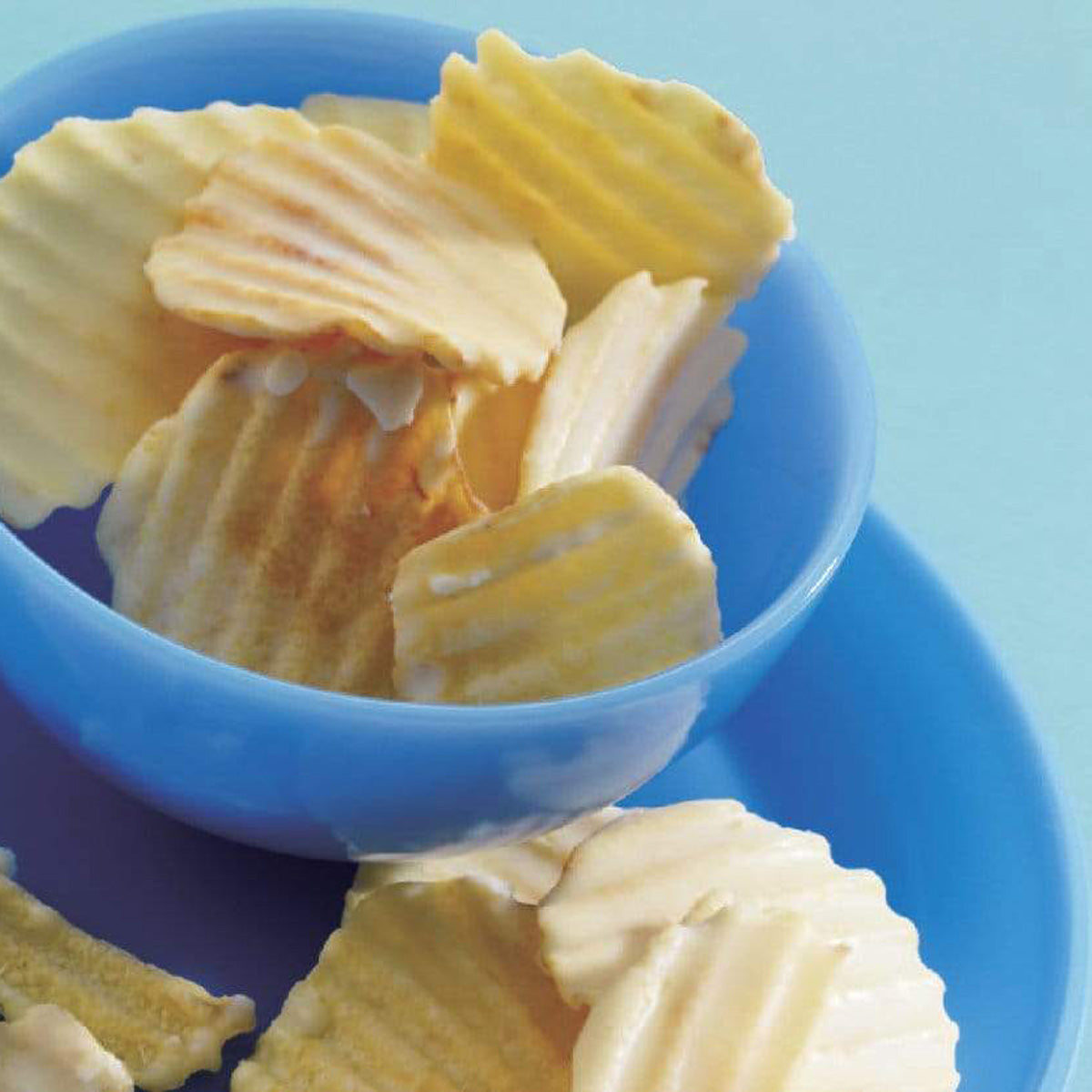 ROYCE' Chocolate - Potatochip Chocolate "Fromage Blanc" - Image shows white chocolate-coated potato chips with a ridged texture inside a blue bowl and plate under. Background is in the color blue.