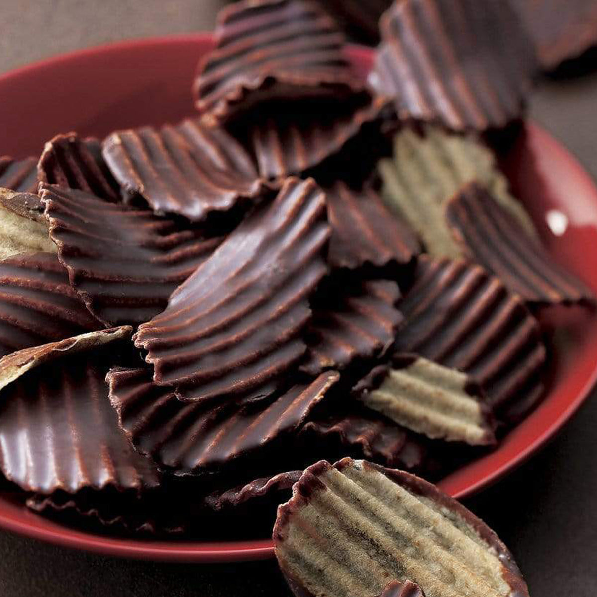 ROYCE' Chocolate - Potatochip Chocolate "Mild Bitter" - Image shows dark brown chocolate-covered potato chips with a ridged texture, as seen in a red plate. Background is in brown color.