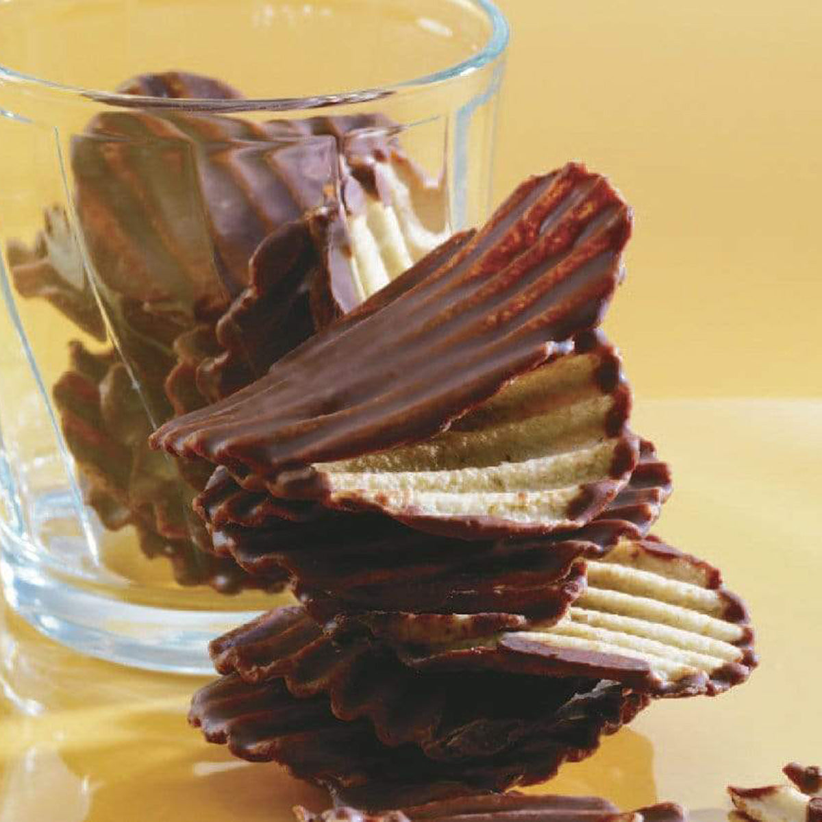 ROYCE' Chocolate - Potatochip Chocolate "Original" - Image shows brown chocolate-covered potato chips with a ridged texture, some on a yellow surface, others in a clear glass behind. Background is in color yellow.