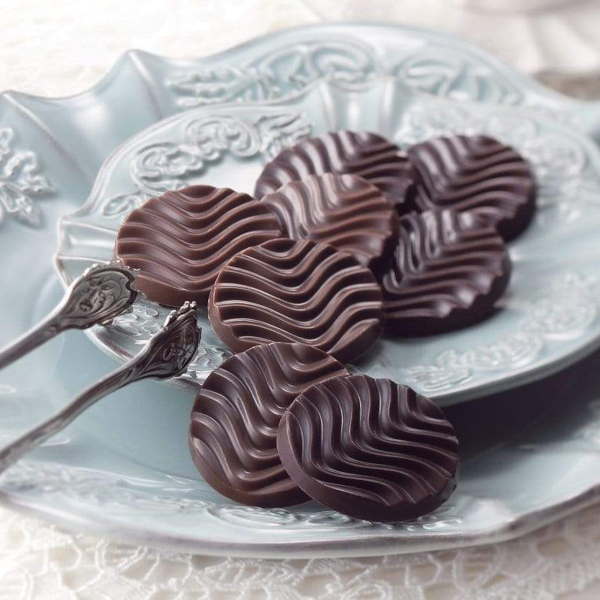 ROYCE' Chocolate - Pure Chocolate "Sweet & Milk" - Image shows brown chocolate discs with a waved texture placed on two light blue plates with engravings. Accents include a silver tong and a white lace table cloth. Background is in white color.