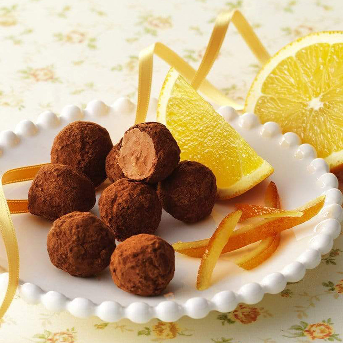 ROYCE' Chocolate - Petite Truffe "Orange" - Image shows brown chocolate balls on a white plate with orange peels and slices. Accents include a gold ribbon and a table background with yellow and green floral prints.