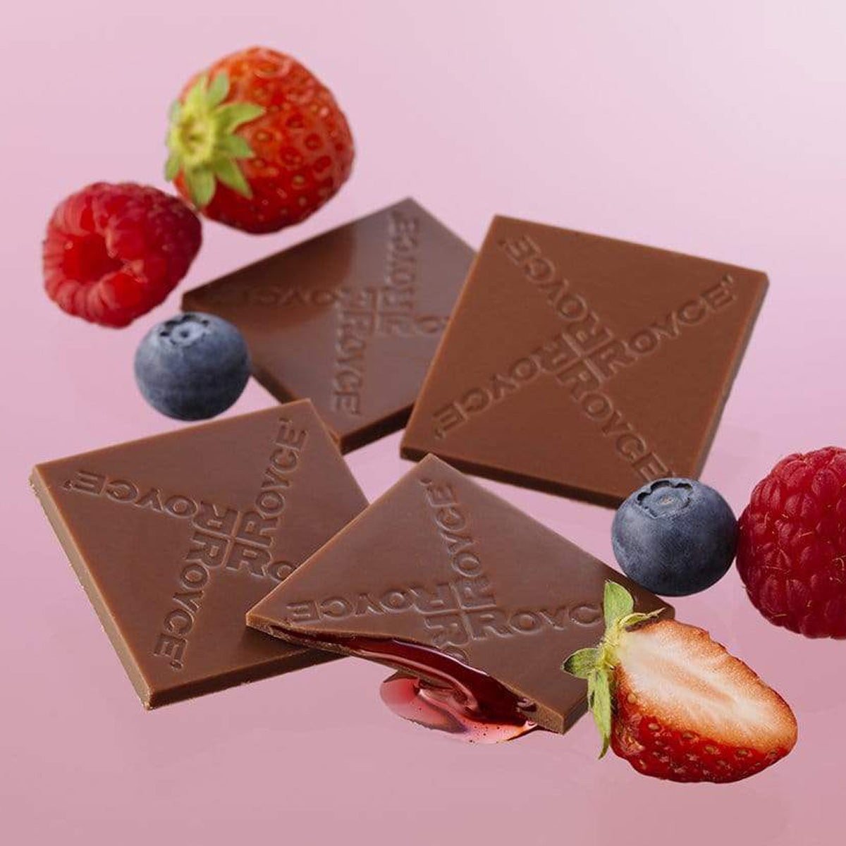 ROYCE' Chocolate - Prafeuille Chocolat "Berry Cube" - Image shows brown chocolate squares with red sauce and engraved with the words "ROYCE'". Accents include red strawberries and raspberries and dark blue blueberries. Background is in pink color. 