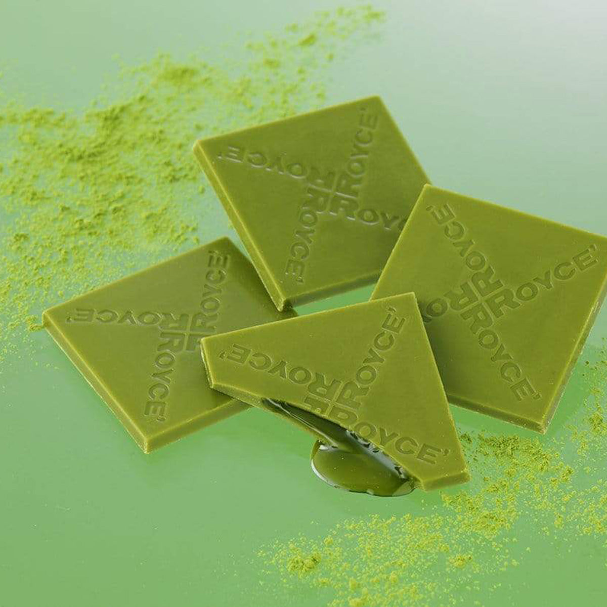ROYCE' Chocolate - Prafeuille Chocolat "Matcha" - Image shows green chocolate squares filled with green sauce and engraved with the words "ROYCE'". Accents include green tea powder scattered. Background is in green color.