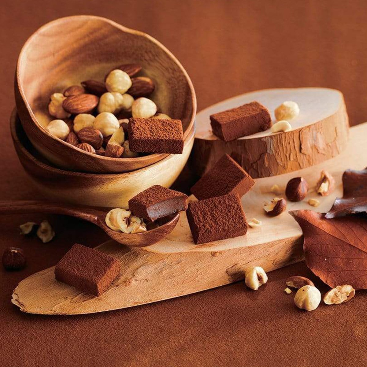 Image shows brown chocolate blocks with loose mixed nuts. Accents include brown wooden bowls and trays together with a brown wooden spoon and some brown leaves. Background surface is in bright brown.