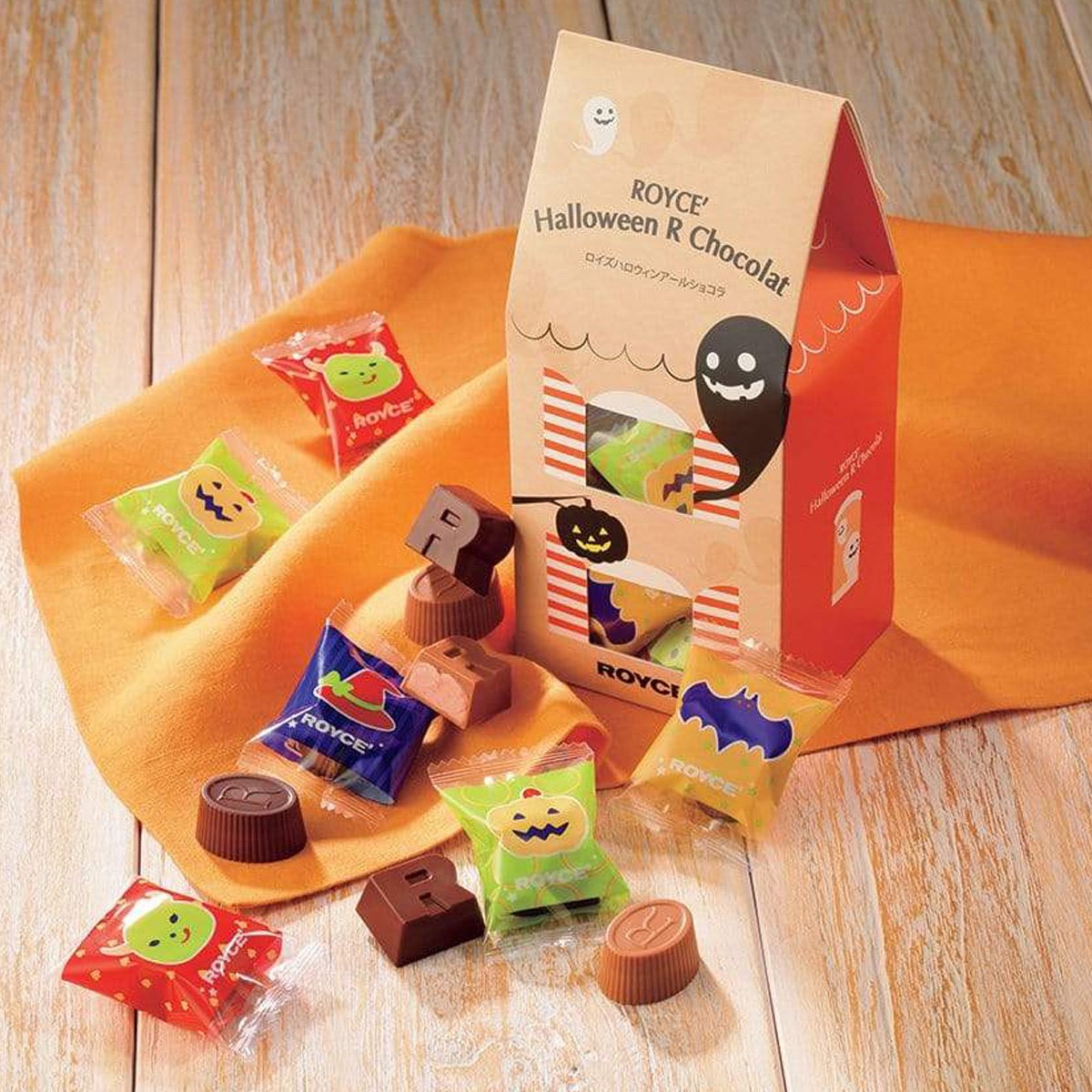 Image shows a box in orange and light brown colors with illustrations of ghosts and pumpkins. Text says ROYCE' Halloween R Chocolat ROYCE'. Accents include R-shaped and oval-shaped chocolates in hues of brown, candy wrappers in red, green, blue, and yellow with prints of pumpkins, ghouls, and a bat. There's also an orange placemat and a brown wooden background.