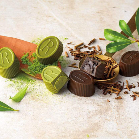 ROYCE' Chocolate - Tea Chocolat "Hojicha & Matcha" - Image shows brown and green chocolate cups engraved with the letter R. Accents include wooden cups, green leaves and powder, and brown tea shavings. Background and surface is light brown.