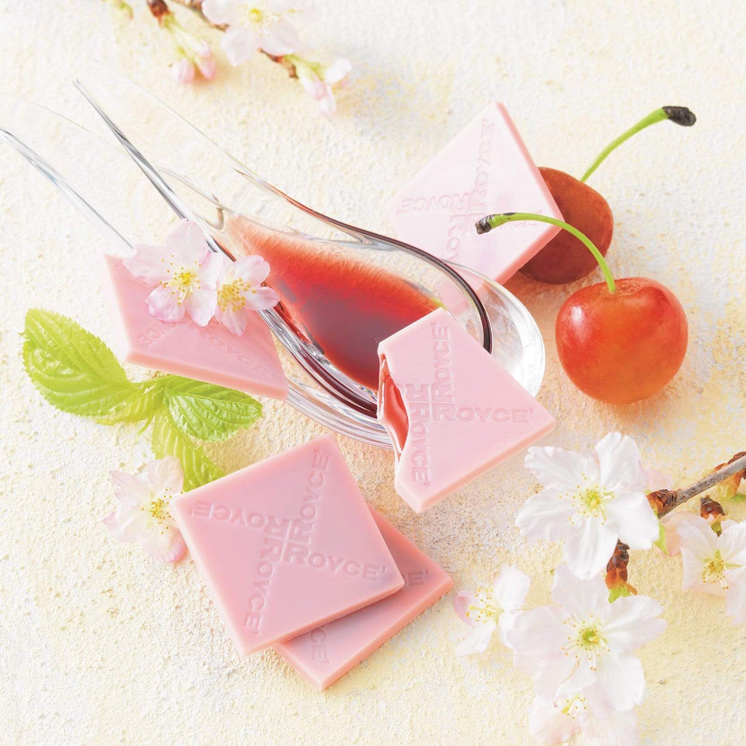 ROYCE' Chocolate - Prafeuille Chocolat "Sakura Cube" - Image shows pink chocolate squares filled with red sauce and engraved with the words "ROYCE'". Accents include clear spoons with red sauce inside, red cherries with green stems, green leaves, and white flowers with brown and gray stems. Background is in color white.