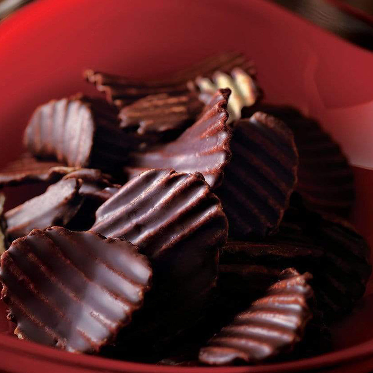 ROYCE' Chocolate - Potatochip Chocolate "Mild Bitter" - Image shows dark brown chocolate-covered potato chips with a ridged texture, as seen in a red plate. Background is in red color.