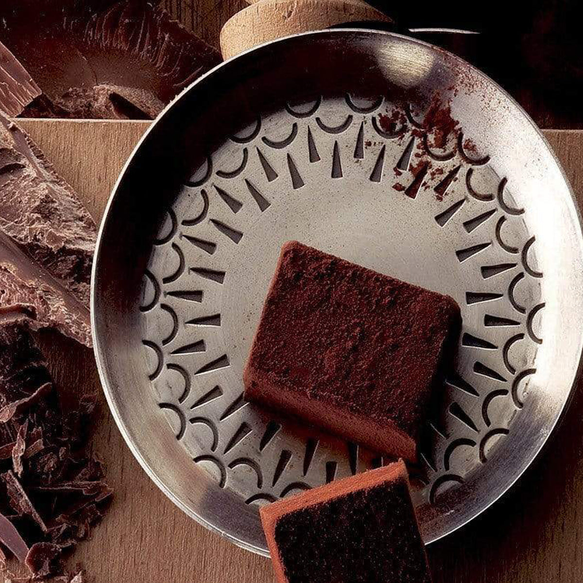 ROYCE' Chocolate - Nama Chocolate "Mild Cacao" - Image shows brown blocks of chocolates on a silver metallic plate. Accents include a brown wooden platform under and brown chocolate shavings on the side.