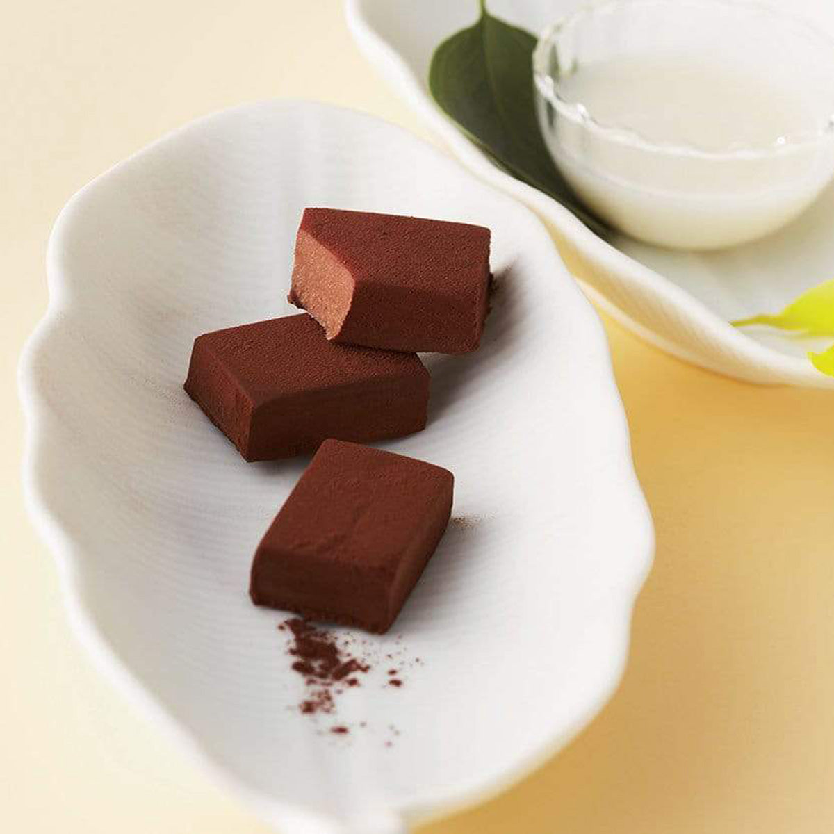 ROYCE' Chocolate - Nama Chocolate "Mild Cacao" - Image shows brown blocks of chocolates on a white leaf-shaped plate. Accents include another white leaf-shaped plate with a bowl of milk and green leaves. Background is in light brown.