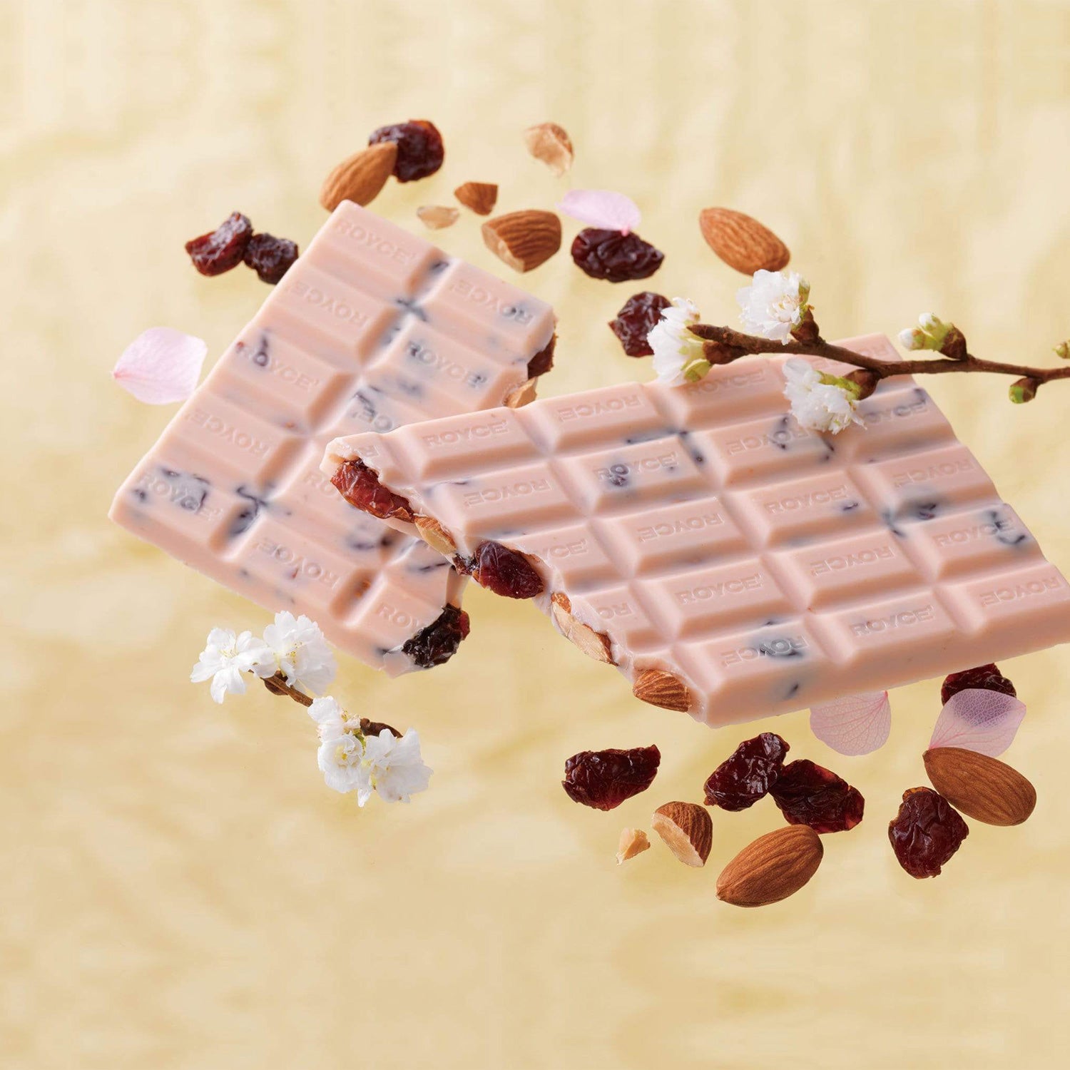 ROYCE' Chocolate - Chocolate Bar "Sakura Berry (Cherry & Almond)" - Image shows pink chocolate bars with dried cherries and almonds, as engraved with the word "ROYCE'". Accents include flowers in white and light pink with twigs in green and brown. Background is yellow in color.