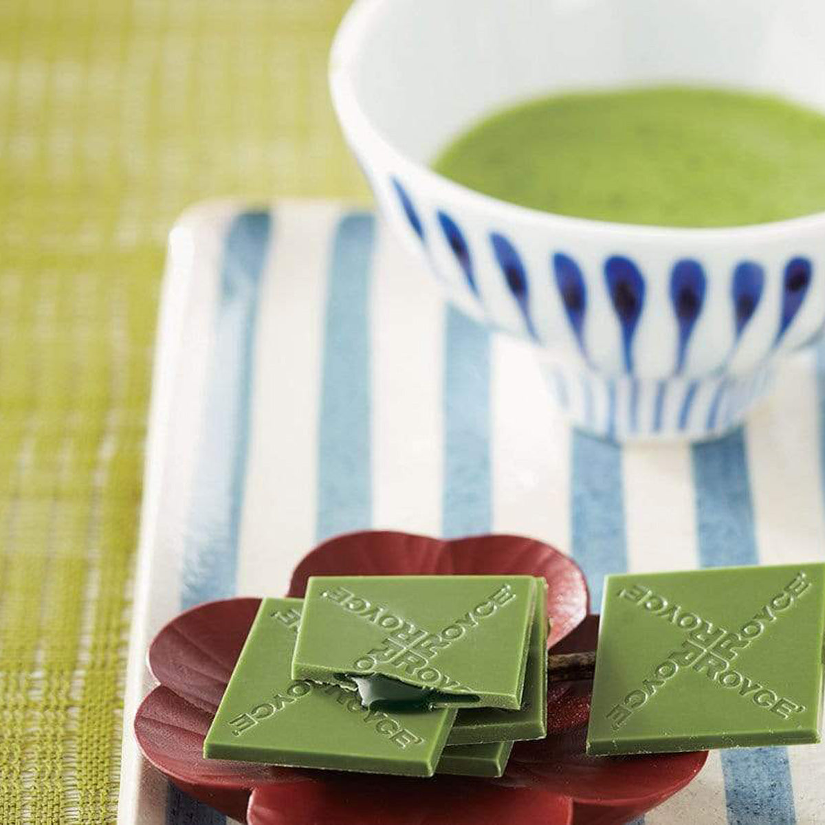 ROYCE' Chocolate - Prafeuille Chocolat "Matcha" - Image shows green chocolate squares filled with green sauce and engraved with the words "ROYCE'", as placed on a brown flower-shaped plate and a plate with blue and white stripes. Accents include a tea cup in blue and white filled with green tea. Background is in green color.
