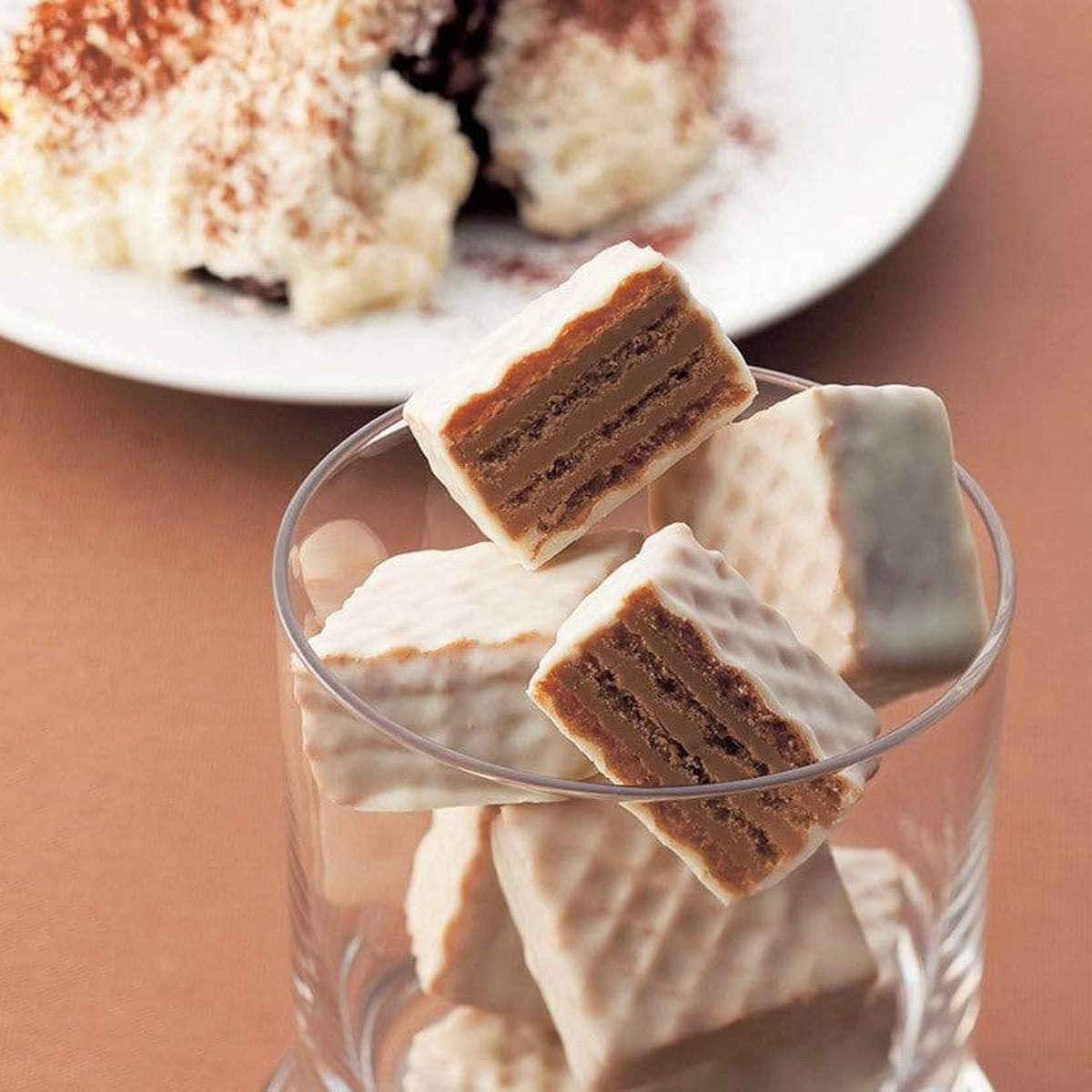 ROYCE' Chocolate - Chocolate Wafers "Tiramisu Cream" - Image shows white chocolate wafers with brown filling inside a clear round glass. Background is in brown color and has a blurred background image of a white plate with tiramisu.